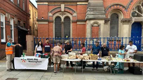 Sewa Day Nottingham Large group of volunteers manning four tables with lots of food for distribution. Man on left learning over a big urn. Food includes sandwiches, bananas and hot drinks.