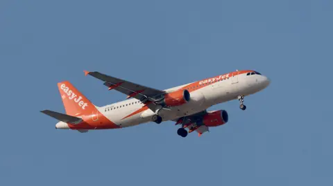 An orange-and-white coloured Easyjet plane in the sky, pictured from below.
