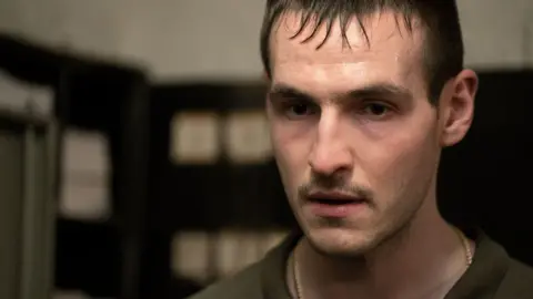 A man with short dark hair and fridge, with a pale complexation is wearing a green top and a chain necklace in the picture. He has a dark moustache. The background behind him is blurred. 