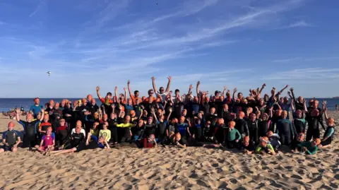A large group of children and adult volunteers with their arms in the air. They're all on the sand at Branksome Chine beach with the sea behind them. They're wearing black wetsuits and orange swimcaps.