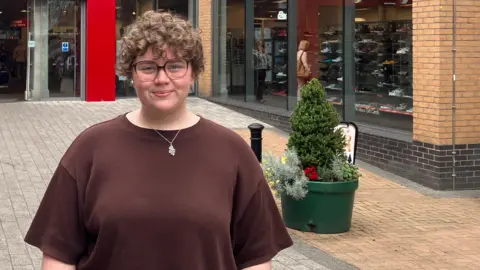 A young woman stands smiling into the camera. Behind her are plate glass windows and a shop doorway. A turquoise planter is behind her left shoulder and contains a small conifer type plant with smaller plants of different colours planted around its base. the young woman has brown curly hair and is wearing glasses with brown frames, a brown t-shirt and a silver pendant.