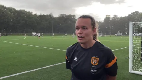 BBC Sophie stands on a rainy football pitch besides the goal.