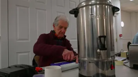 Bernard Crick, who has light grey hair and wears a red fleece, sits at a table repairing an electrical item. 