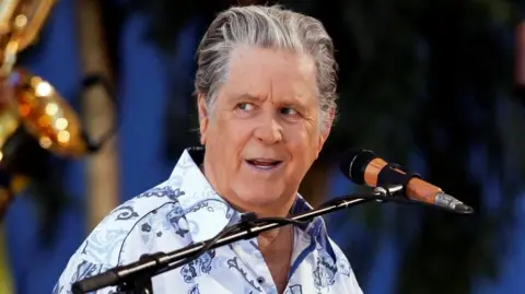 Brian Wilson appears on stage.  He is wearing a white, patterned shirt and is in front of a microphone. 