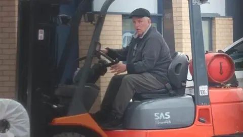 A man wearing a flat cap and a grey cardigan and grey pants sits on top of a forklift truck which is holding a rolled tube of artificial grass. 