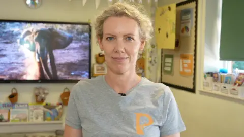Qays Najm/BBC A woman with blonde curly hair that is tied up smiles at the camera. She is inside a classroom and wears a grey T-shirt. There is a picture of an elephant on a screen behind her. 