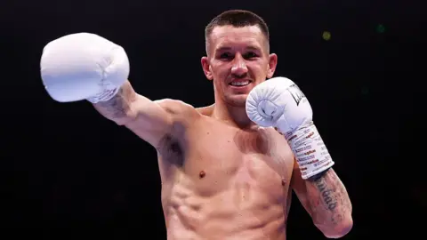Getty Images A shirtless man with short brown hair is smiling and is mid punching the air in a forwards motion. He is wearing white boxing gloves