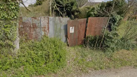 Rusty, corrugated metal fencing across the edge of a field with part of a road visible in front of it. The fencing is surrounded by trees and shrubbery.