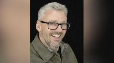 Craig Hurcombe smiles at the camera. He wears a green collared jacket and black rimmed glasses. He has short grey hair and a beard. 