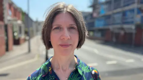 A close up picture of Haf smiling looking at the camera. She is standing on a pavement with a road and buildings behind her. Haf is wearing a multicoloured patterned shirt. She has short brown hair. It is a head and shoulders shot of her.