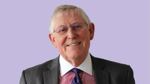 Close up of Peter Burroughs. Mr Burroughs is wearing a blazer with a red and white striped shirt and a navy tie. He is looking into the camera and smiling.