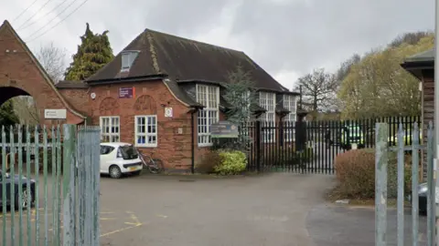 A single storey brown brick library building, in front of which is a small car park and metal fencing