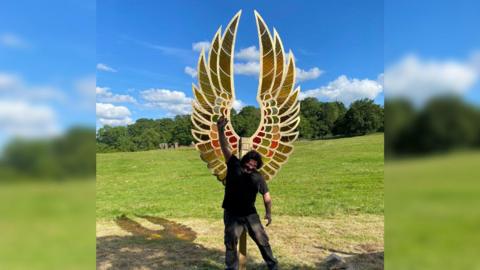 Edgar Phillips standing in front of a pair of tall golden pointed wings with his arm raised in a fist
