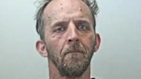 Police custody image of Aaron Cane with brown beard and receding brown hair wearing a grey t-shirt.