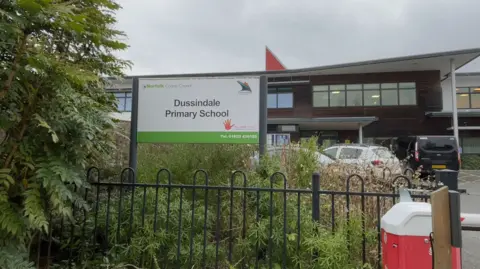 Robby West/BBC The exterior and entrance of Dussindale Primary School. The metal gates, some bushes and a school sign are visible. In the background is the school building, which has a wood-panelled effect.