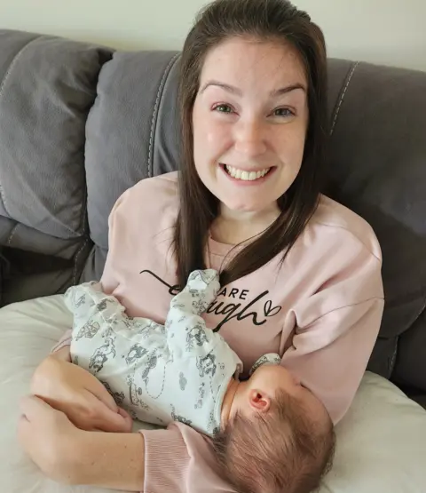 Lucy Lintott Smith Sitting on a grey sofa and wearing a pink jumper, Lucy Lintott Smith beams as she hold her third baby, who is supported on a maternity cushion. 