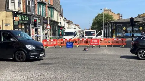 The image shows a junction that has been partially closed off in a town centre with shops, traffic lights, and multiple vehicles.
