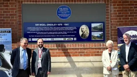 City of Doncaster Council A circular blue plaque reading: "Sir Nigel Gresley, railway engineer, designed the mallard and flying scotsman locomotives". Below it is a larger, navy plaque with more biographical information. Three men and a woman stand in front. The woman is Mayor Ros Jones, who has short white hair and wears a white suit.