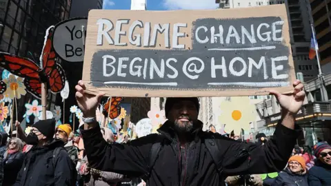 Getty Images A man holds a sign that says "regime change begins @ home"