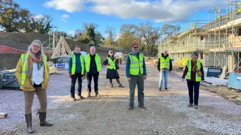 Joanna stands to the left on a road in the Clay Park building site, with trustees of the Transition Homes Community Land Trust standing to her right, all wearing high-viz jackets. The sky is blue with white clouds, while to the right the timber frames of the new social housing can be seen in construction.
