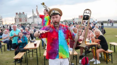 Durham County Council A saxophone player from Mr Wilson's Second Liners performs among the crowd at Durham Brass Festival in 2025. He is wearing a hat and colourful shirt. People sitting on benches behind him are smiling and clapping. 