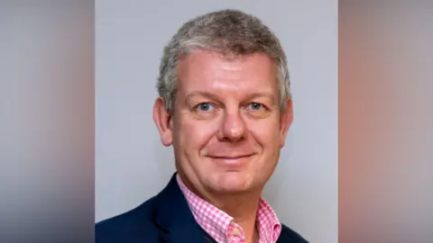 DLN ICB Cluster A head and shoulders shot of a man with grey hair and a pink and white shirt. He is smiling into the camera. There is a grey background.