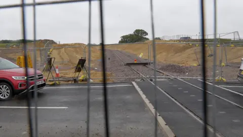 Richard Daniel/BBC View through a metal fence shows a road bisecting a rail line that is under construction. A red car can be seen at the left of the frame.