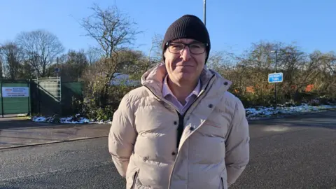 Ian Hudson is wearing a cream coat with a black woollen hat. behind his is the proposed restaurant entrance.  