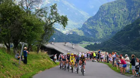 Getty group of cyclists racing along a winding mountain road surrounded by lush green hills and towering peaks. Spectators line both sides of the road, cheering and taking photos under a bright blue sky. The cyclists wear colourful jerseys and helmets, with one in yellow leading the pack, creating a lively and energetic scene in a picturesque alpine setting.