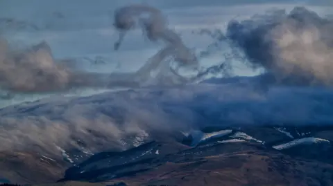 BBC Weather Watchers / Ronaldo Grey clouds make swirling patterns in the sky, above a much larger bank of cloud hugging the top and sides of a mountain range in Cumbria. Beneath the clouds, the fells are topped with a dusting of snow.