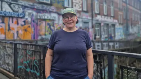 Zoe Genders wearing a green cap and dark blue T-shirt stands on a metal bridge railing in front of a large brick building covered in colorful graffiti and murals. The background is softly blurred, showing windows, street art, and a canal or waterway running alongside the building. She is wearing glasses.