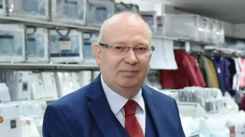 Kevin Baldwin wearing glasses and navy suit with a white shirt and red tie. He is standing in one of his store in front of two beds and shelves filled with bedding items. He is looking into the camera.