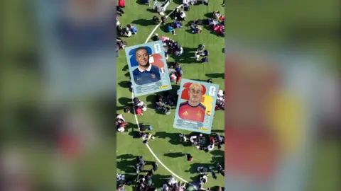 Scores of people gathered on a football pitch, with World Cup stickers of Kylian Mbappé and Lamine Yamal superimposed.