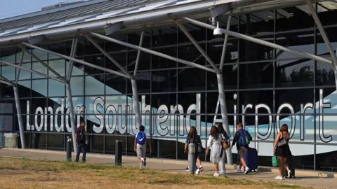 Passengers outside the terminal building at Southend Airport. the building is a mostly glass structure. People wear summer clothes and it is sunny. 
