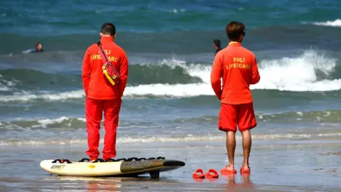 Two RNLI lifeguards stood on a beach, both looking towards the sea where there are people swimming. They are dressed in red branded clothing with RNLI lifeguard clearly written on their backs. There is a yellow surfboard on the ground along with some red sandals. 