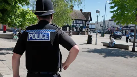The back of PC Byrne as he walks down Shirley high street on a sunny day. He is wearing black with Police written across his back in white letters. He is walking across a paved square surrounded by shop. There are people sitting on benches and three trees.