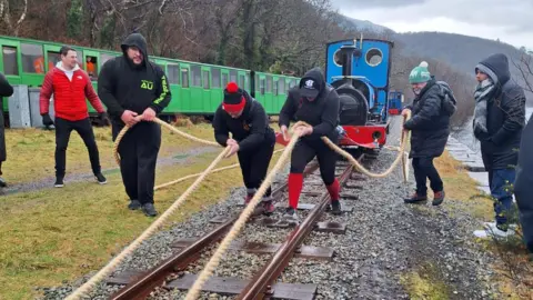 Sue Taylor-Franklin Sue and Sam are holding ropes as they pull a red, blue and black train behind them. They are wearing black and red clothes and pulling the train on a train track. They are people on either side of them also holding the rope to assist them as they pull. 