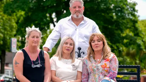 PA Media Three middle aged women sit on a black bench looking into the camera with serious expressions. Standing behind the bench is a middle aged man with grey hair, a grey beard and a white shirt. 