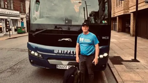 Supplied Alan stands in front of a bus on a street. He wears a blue shirt and a black baseball cap.