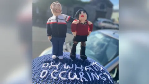 A postbox topper showing a knitted version of Gavin and Stacey with the famous catchphrase used by Nessa, played by Ruth Jones, of "what's occurring?",  stitched in white on a blue base.