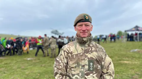 Col Matt Palmer stood to the right looking at the camera. There are crowds of people stood on the grass with a helicopter taking off in the background.