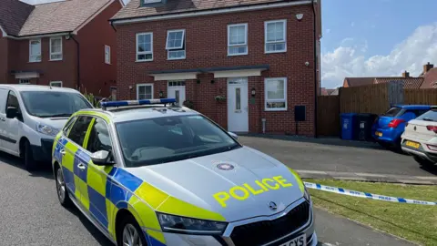 A police car outside a three-storey new build semi-detached house with white windows and door. There is police tape outside the Tarmac'd area in front of the house. Two cars are parked on this.