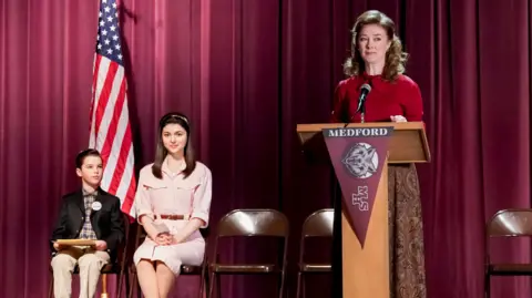 Getty Images Mahaffey stands at a podium on the set of young sheldon. she is wearing a red sweater and brown skirt. to the side, a young girl and young boy sit on folding chairs. there is a purple curtain behind them and an american flag.