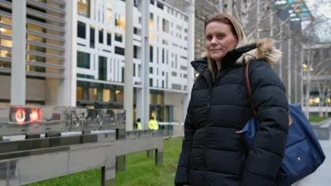 Micala, who has long blonde hair and is wearing a thick padded black coat, stands outside a large office block with a sign saying Home Office outside.