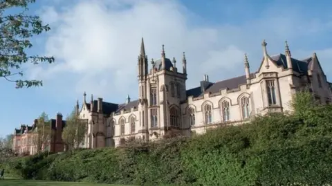 UU The university campus in Londonderry. A number of bushes and grassy areas can be seen in front of a Gothic-looking building with several large windows.