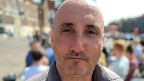 A picture of Jason smiling with his mouth closed at the camera. He is bald with light stubble on his face, and is outside in front of what appears to be a group of people sitting around tables at a bar or cafe. 