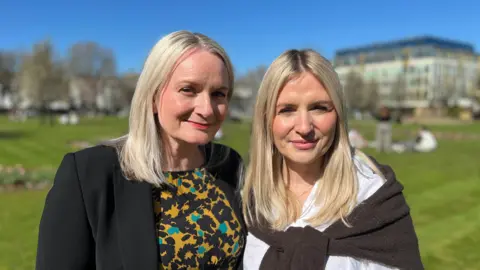 Helen and Georgina stand side by side outside. They are standing on grass with a few people sitting behind them. They both have medium-length blonde hair and are smartly dressed.