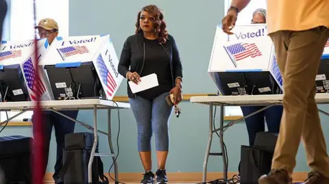 People casts ballots during early voting at the Garners Ferry Adult Activity Center on 26 May 2026 in Hopkins, South Carolina. A woman in a black top and gray leggings walks between two voting tables, holding a white piece of paper and her phone