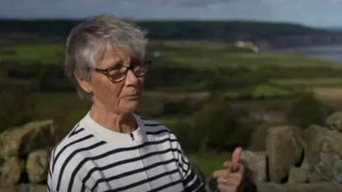 Phil Bodmer/ BBC Margaret Atkinson stood outside, overlooking the coast