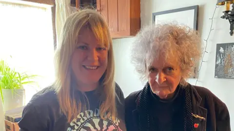 A woman with long blonde hair and a fringe wearing a dark grey T-shirt stands next to an older woman who has short, curly grey hair and is wearing a brown jacket. They are both smiling at the camera.